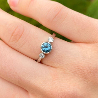 Platinum teal sapphire and diamond trilogy engagement ring shown worn on hand.