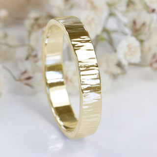 A 18ct gold wedding ring with a hammered tree bark texture, displayed against a floral background.