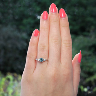 Fair trade teal sapphire and diamond trilogy ring shown on the hand with greenery in the background.